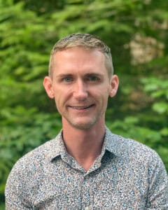 Smiling short haired man in pale blue sweater standing in front of trees - James Sullivan Orchard Recovery Center Registered Nurse
