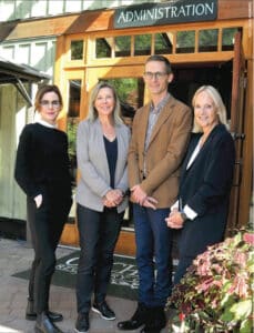 3 smiling women and 1 smiling man standing in front of a wooden building with the sign Administration over the door