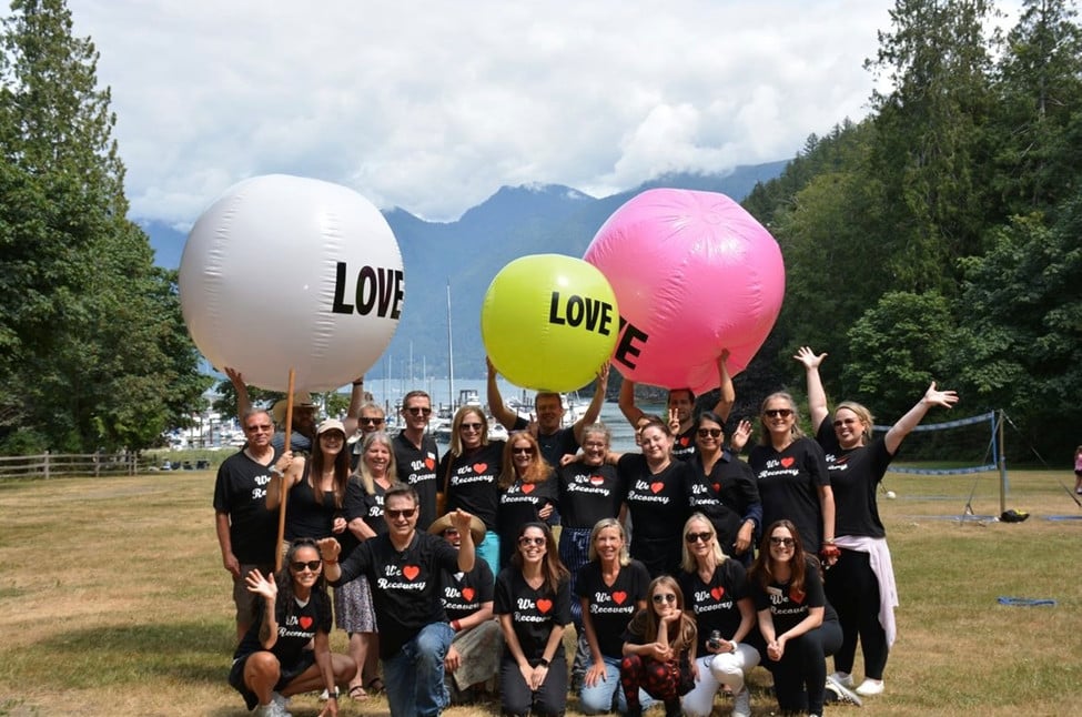 Group of men and women wearing t-shirts with the words "We Love Recovery". Several of the people are holding up 3 large balls with the word Love on them. The balls are white, yellow and pink. In the background is a volleyball net on a field in front of a small marina. Mountains with clouds on top are across the water.