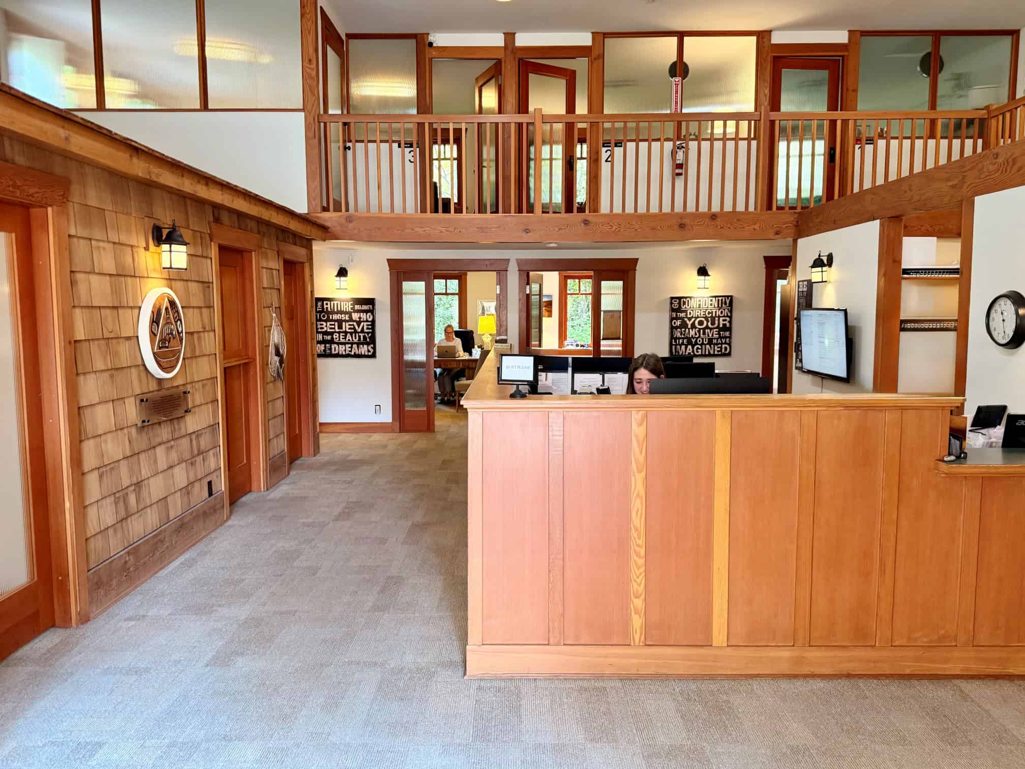 Two level office with wooden partition. A young woman with black hair is sitting at a computer. In the distance is a blonde lady working at a computer in a separate office. Several offices are on the 2nd floor with a wooden railing. Native art is on the left wall. Administration office Orchard Recovery Center.