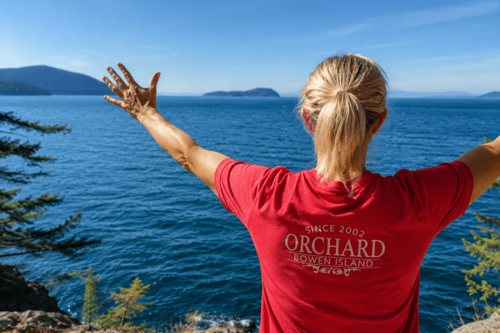 Woman with a blonde ponytail looking out over an ocean to land in the distance. She is wearing a red t-shirt from Orchard Recovery Center