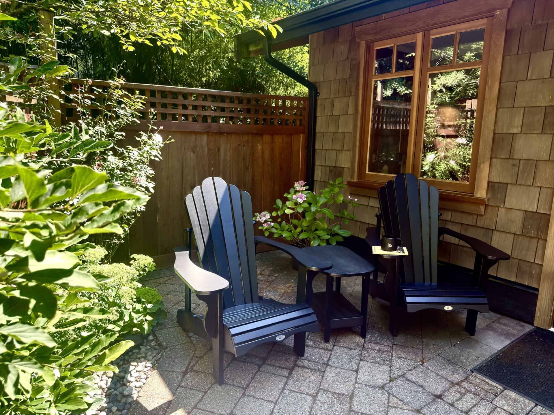 Two blue wodden chairs with a small blue table in between them. A pad of paper and coffee mug are on one of the arms of the chair. The wall of a building with wooden shakes and a double window are on one side. A wooden fence is on another side and bushes are on the third side.