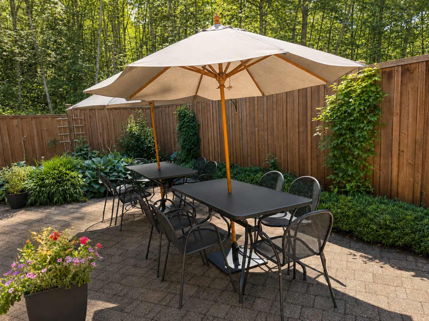 Two metal tables and chairs both with patio umbrellas on a stone patio area. Wooden fence with bushes and flowers surround the area.