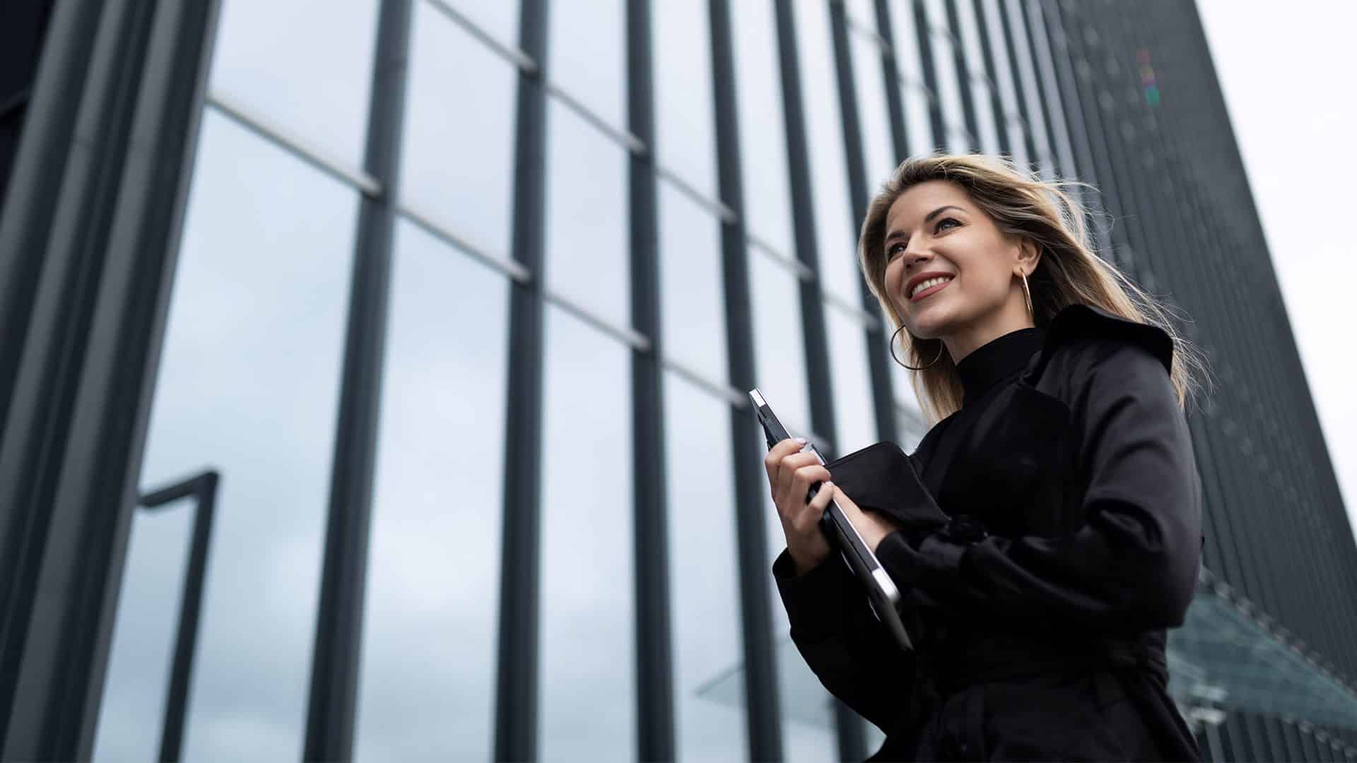 Professional woman standing outside modern office building, smiling, holding a tablet and smartphone, showcasing success and business growth.