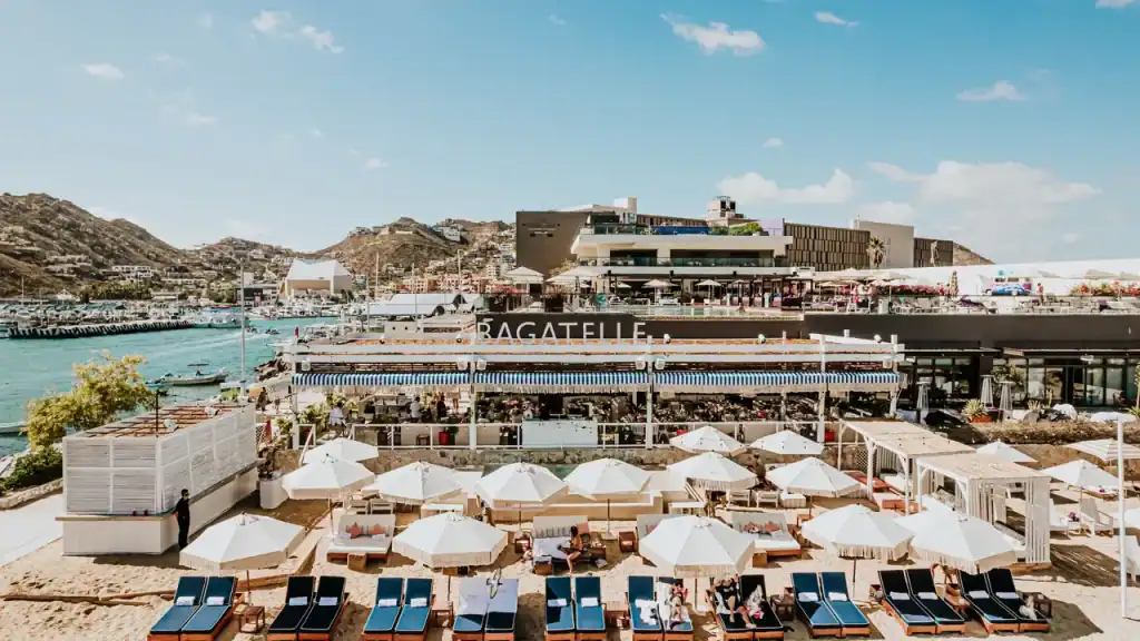 Vibrant Palmilla Waterfront Beach Scene With Lounge Chairs, Umbrellas, And A Lively Restaurant Overlooking The Marina And Hillside Neighborhoods.