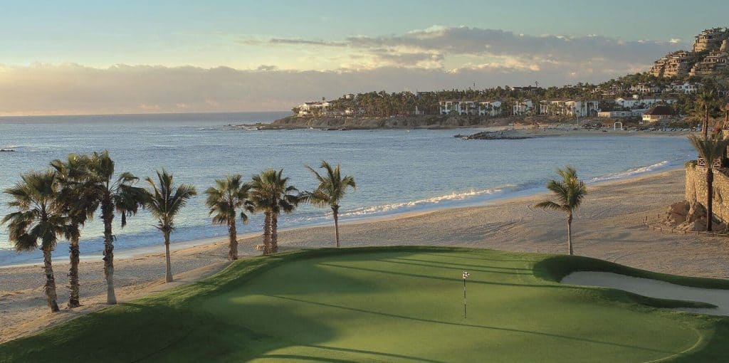 Lush Green Golf Course With A Beach And Ocean View, Palm Trees Lining The Shore, And Upscale Homes On The Hillside, Representing Luxury Real Estate And Resort Living In Palmilla.
