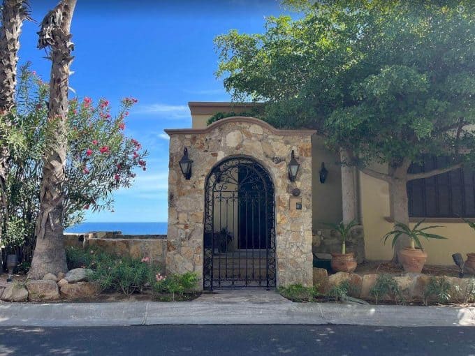 Luxurious Palmilla Gated Entrance With Stone Facade, Black Wrought Iron Gate, Lush Greenery, And Ocean Views In The Background, Showcasing Elegant Tropical Home Design.