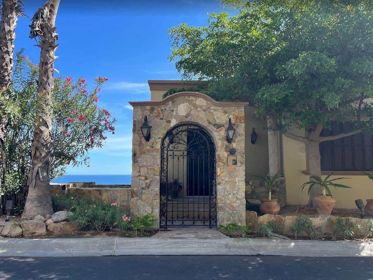 Luxurious Palmilla Gated Entrance With Stone Facade, Black Wrought Iron Gate, Lush Greenery, And Ocean Views In The Background, Showcasing Elegant Tropical Home Design.