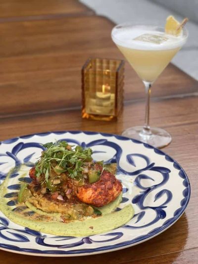 Juicy Grilled Chicken Topped With Fresh Herbs And Vegetables, Served On A Decorative Blue And White Plate With A Creamy Green Sauce, With A Refreshing Lemon Cocktail And Candle On A Wooden Table At Palmilla.