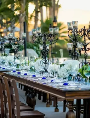 Elegant Palmilla Wedding Reception Setup With A Long Wooden Table, Floral Centerpieces, And Intricate Candelabras In A Scenic Outdoor Setting During Sunset.
