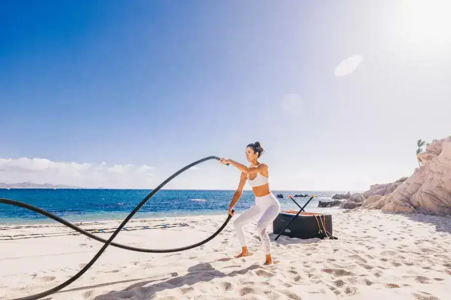 Fitness And Wellness – Homes For Sale In Palmilla Palmilla Beach Scene With A Woman Exercising With Battle Ropes On White Sand Against A Bright Sunny Sky.