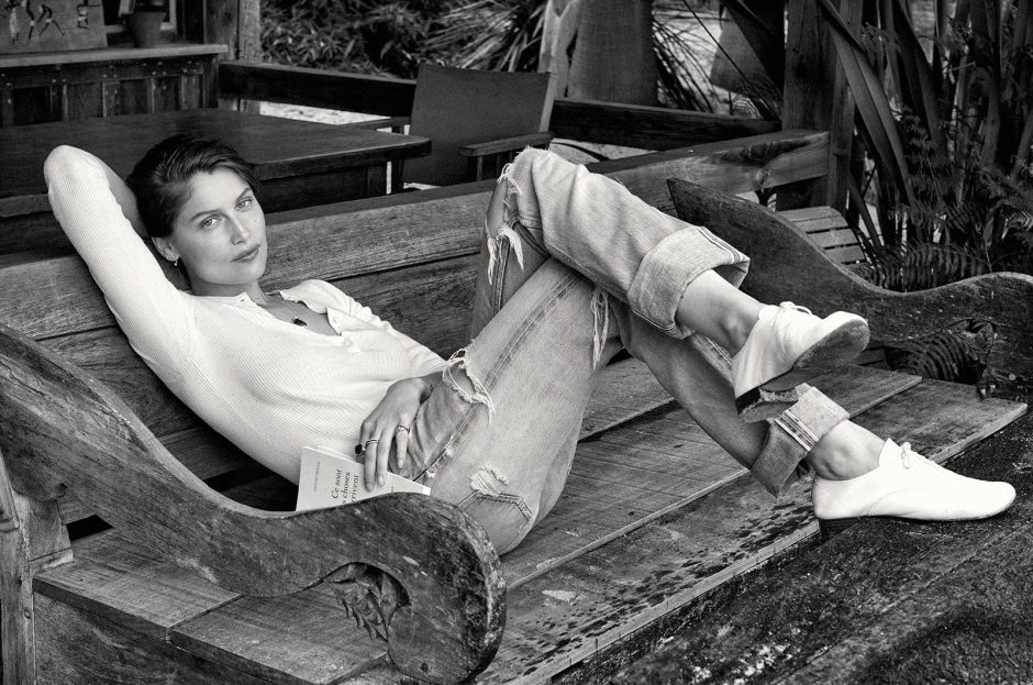 Relaxed woman lounging on rustic wooden bench, black and white portrait, casual denim and white sneakers, outdoors garden setting, stylish and contemplative vibe, high-quality fashion photography.