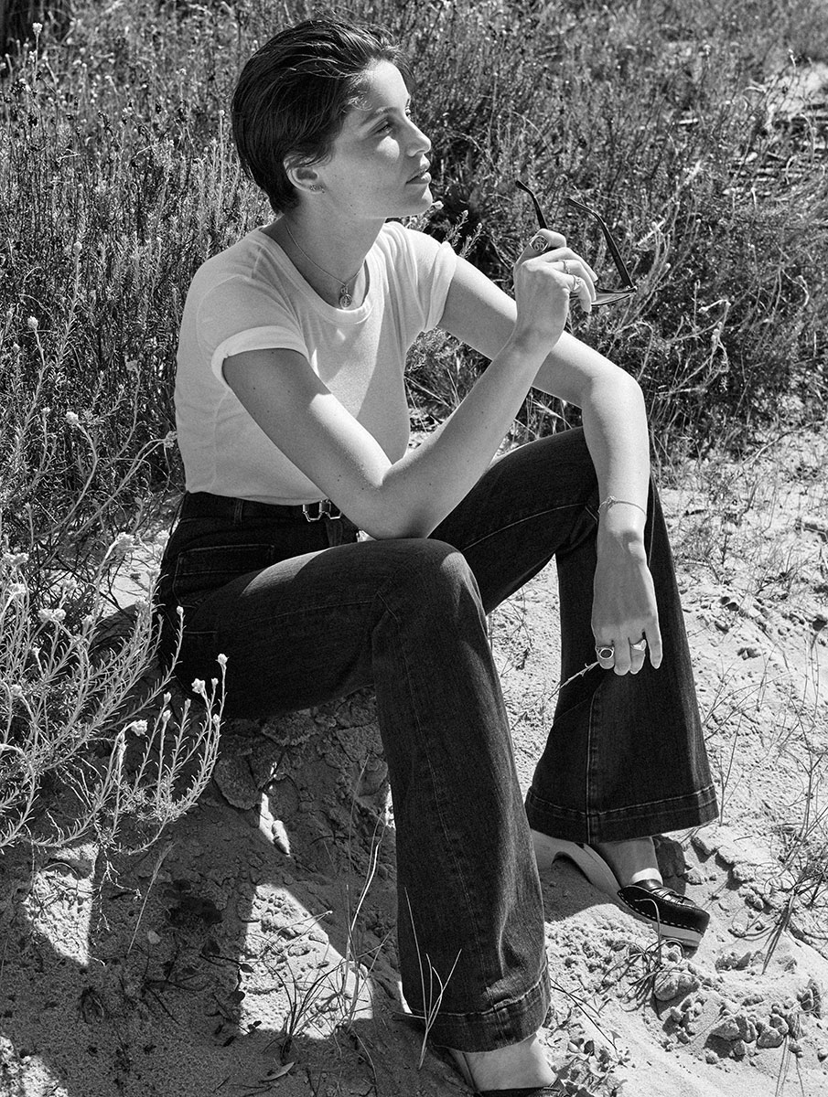 Elegant black and white portrait of a young woman sitting on sandy terrain, gazing into the distance, holding sunglasses, showcasing fine art portrait photography by Pamela Hanson.