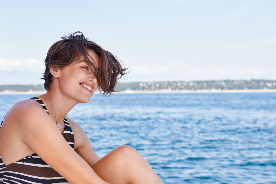 Vibrant portrait of a young woman with short, tousled hair, wearing a striped swimsuit, smiling by the ocean during daytime, showcasing contemporary fashion and natural beauty.