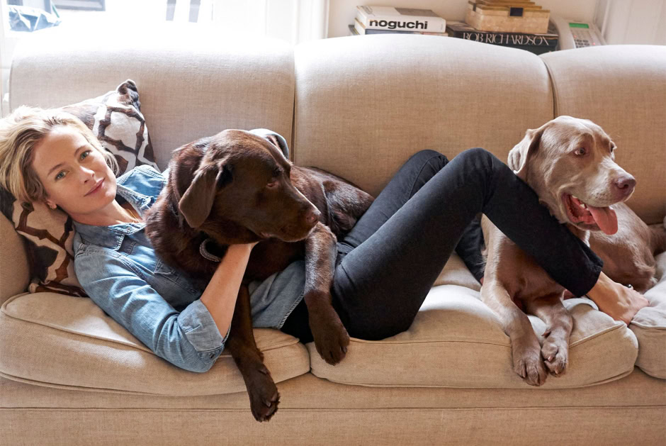 A young woman relaxing on a beige sofa with two large dogs, one brown and one yellow, in a cozy, well-lit living room with books and decorative pillows.