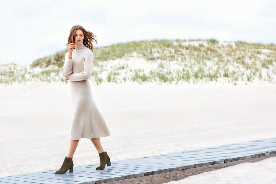 Elegant woman in beige knit dress and ankle boots walking on a wooden pathway in a sandy, beach dune landscape with sparse green vegetation and a cloudy sky.