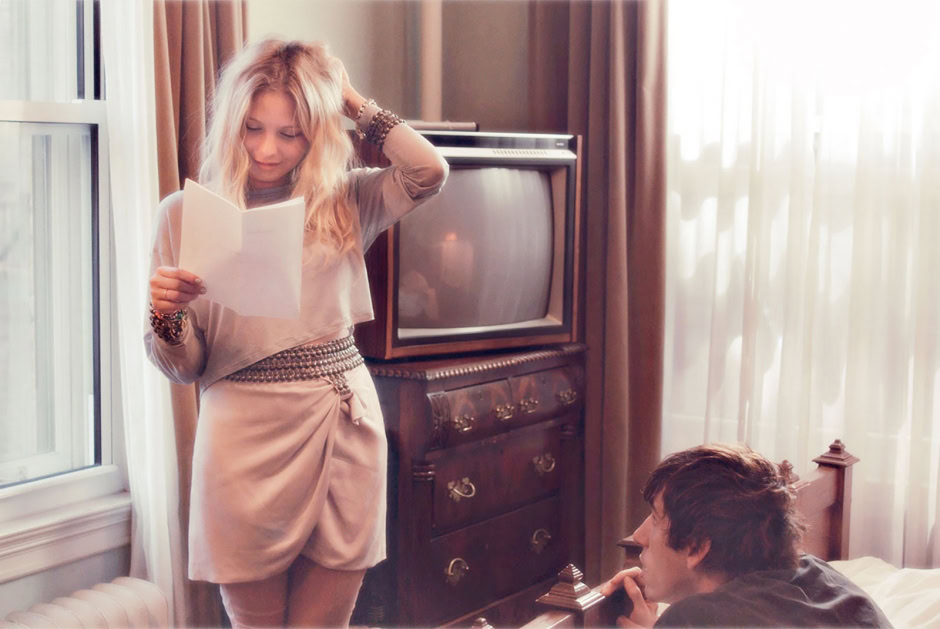 Soft natural light enhances intimate moments in vintage-inspired bedroom scene; young woman reading while standing, young man lying on bed with curious gaze, classic decor, and retro television.