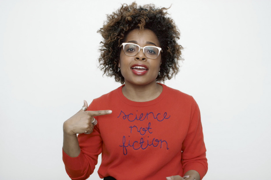 Beautiful woman wearing glasses and a red sweater with "science not fiction" written on it, pointing at her quote, in a professional portrait photography shot emphasizing empowerment and gender equality.