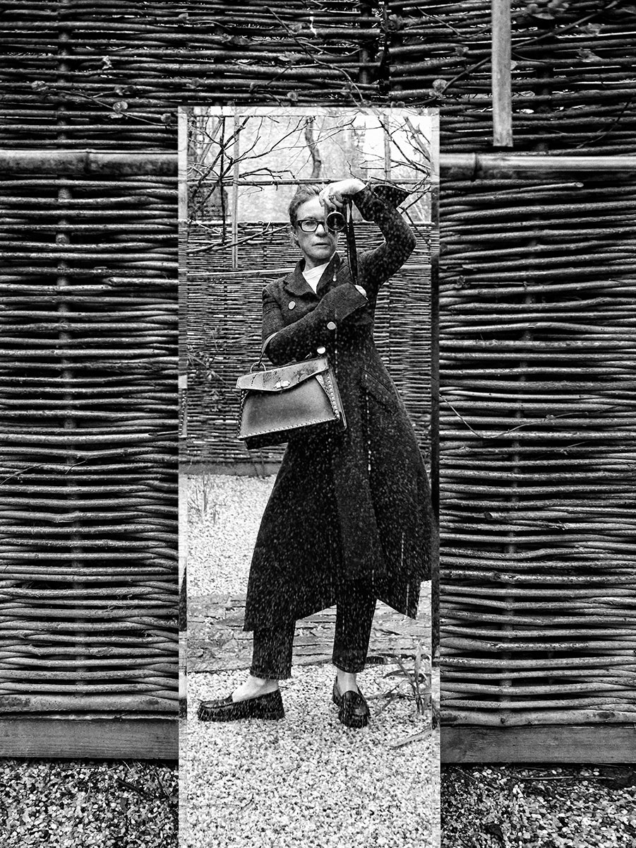 Black and white portrait of a woman taking a selfie with a camera, standing outside behind a woven wooden fence, wearing fashionable clothing, glasses, and carrying a handbag, artistic photography shot by Pamela Hanson.
