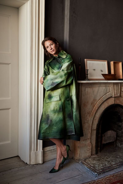 Elegant woman wearing a green leather coat standing beside a fireplace in a stylish indoor setting.