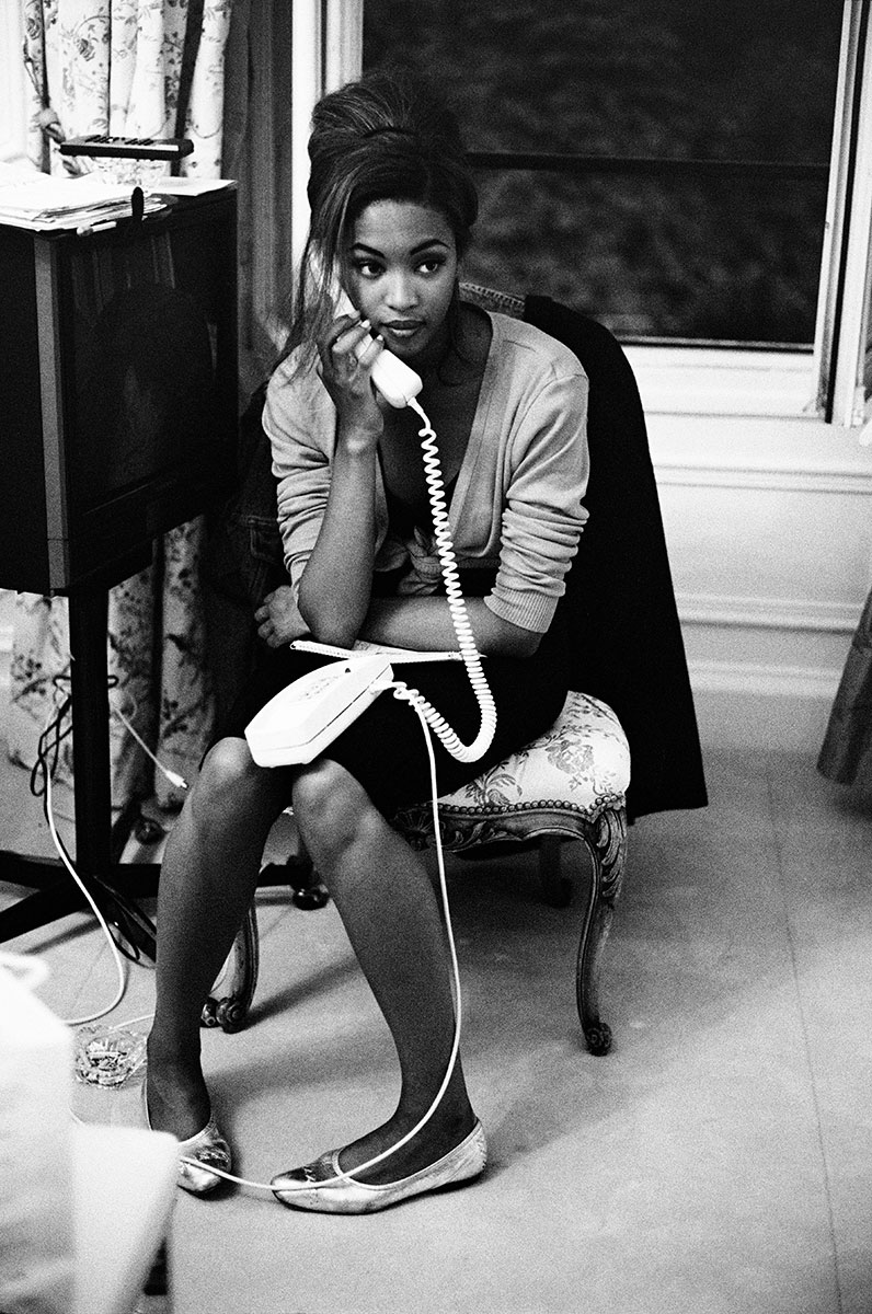 Vintage black and white portrait of a woman with styled hair, sitting with legs crossed, holding a landline phone in a cozy indoor setting, showcasing Pamela Hanson’s elegant fashion photography style.