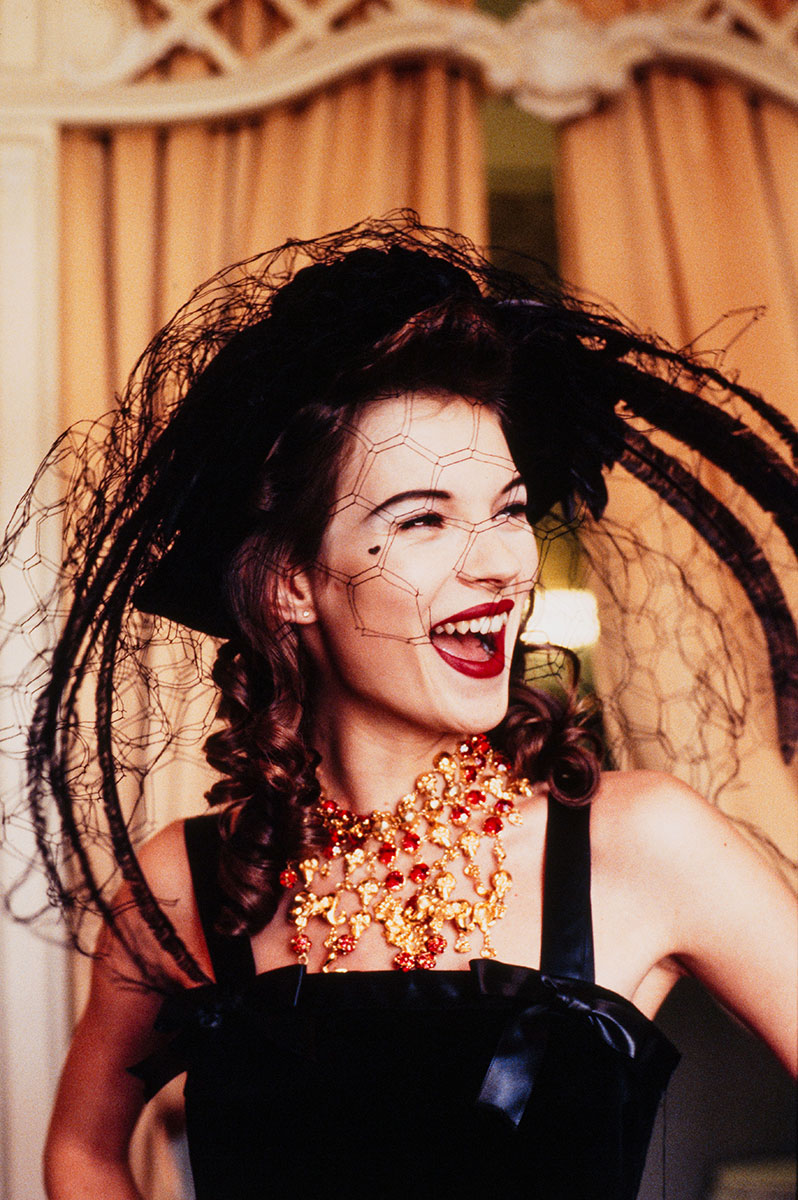 Elegant woman wearing vintage black dress and elaborate jewelry, smiling joyfully at a themed event or photoshoot, with stylish hat and intricate veil for a glamorous, high-fashion look.
