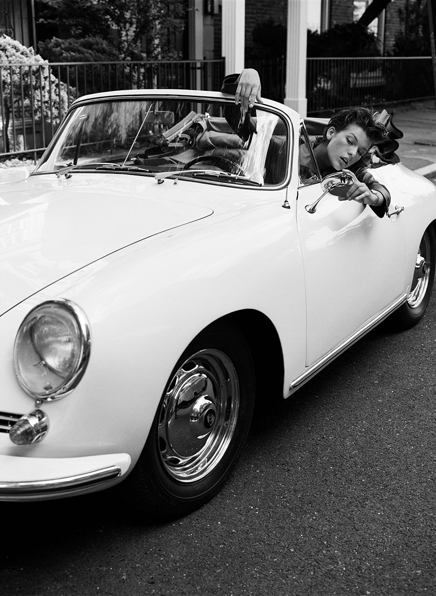 Timeless black and white photo of a woman leaning out of a vintage convertible car, posing playfully with phone in hand, outdoor setting with modern house background, classic cars, fashion photography, lifestyle shot.