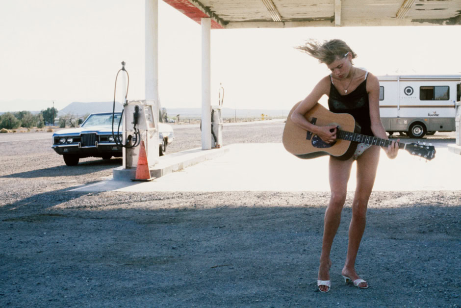 Guitar-playing woman at a vintage desert gas station, captured in a cinematic style, emphasizing Americana and artistic photography.