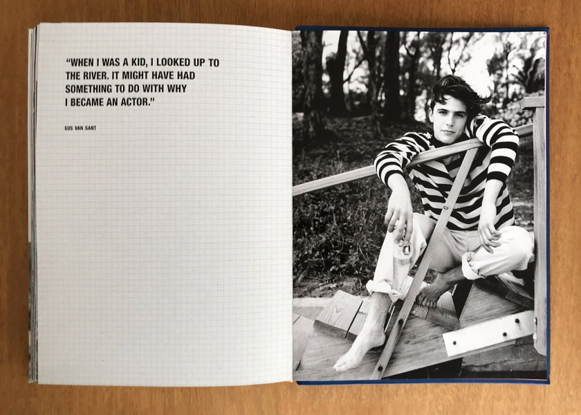 A black and white portrait of a young man sitting outdoors on a wooden structure, wearing a striped shirt, with a natural forest background.