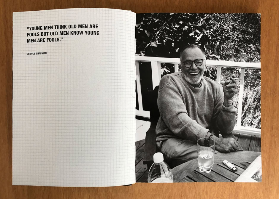 Vintage black and white portrait of a smiling man with glasses, sitting outdoors on a wooden deck with lush foliage in the background, capturing his joyful expression and relaxed vibe.