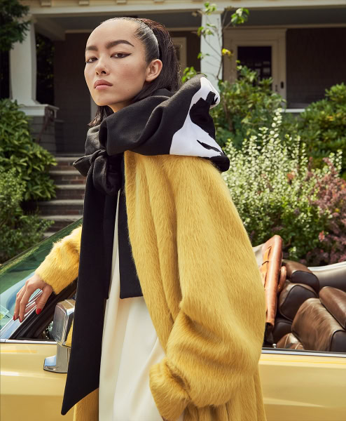 Fashion photography of a stylish woman in a vibrant yellow faux fur coat and graphic hoodie, standing beside a vintage convertible car in a suburban neighborhood, showcasing upscale portrait and contemporary fashion photography.