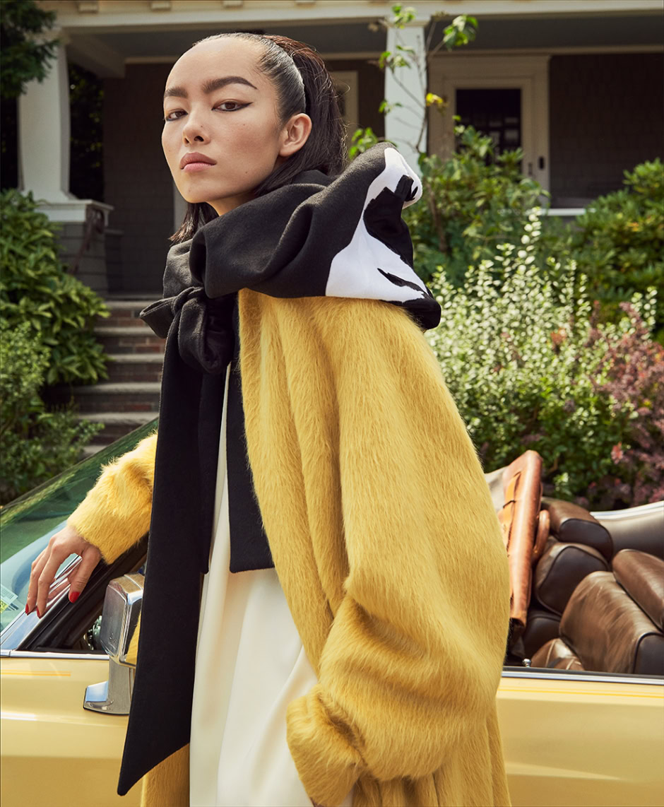 Fashion photography of a stylish woman in a vibrant yellow faux fur coat and graphic hoodie, standing beside a vintage convertible car in a suburban neighborhood, showcasing upscale portrait and contemporary fashion photography.