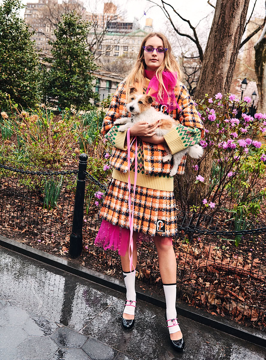 Elegant woman holding a small dog in a colorful outdoor city park during autumn, wearing high-fashion patterned clothing, sunglasses, and stylish shoes for contemporary photography.