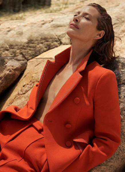 Elegant woman relaxing outdoors in a stylish orange blazer and matching pants, sunlit rocky background, showcasing fashion photography by Pamela Hanson.
