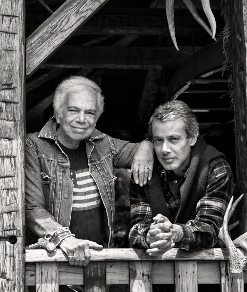 Portrait of two men, one older with white hair and one younger, in black and white, captured in a rustic outdoor setting, showcasing natural light and candid expressions.