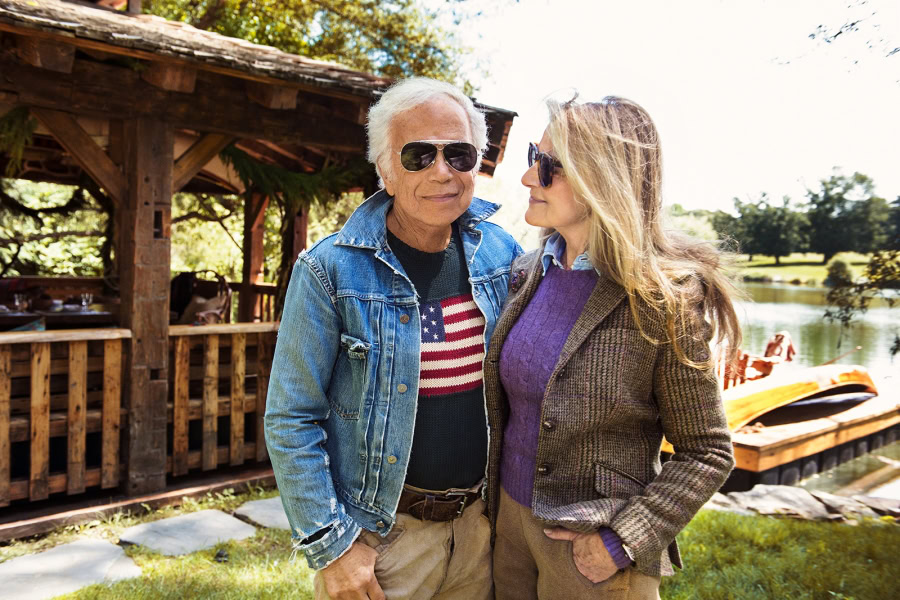 A man and woman wearing sunglasses standing outdoors near a lake with a wooden dock and paddleboat, showcasing lifestyle and portrait photography by Pamela Hanson.