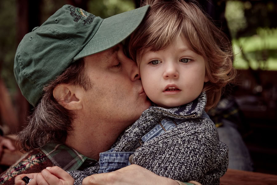 A man wearing a green cap kisses a young boy with curly brown hair, capturing an intimate, candid moment outdoors with natural light.