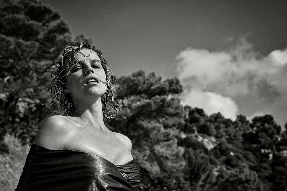 Elegant black and white portrait of a woman with curly wet hair, outdoors with a rocky landscape and cloudy sky in the background, showcasing professional fashion photography.