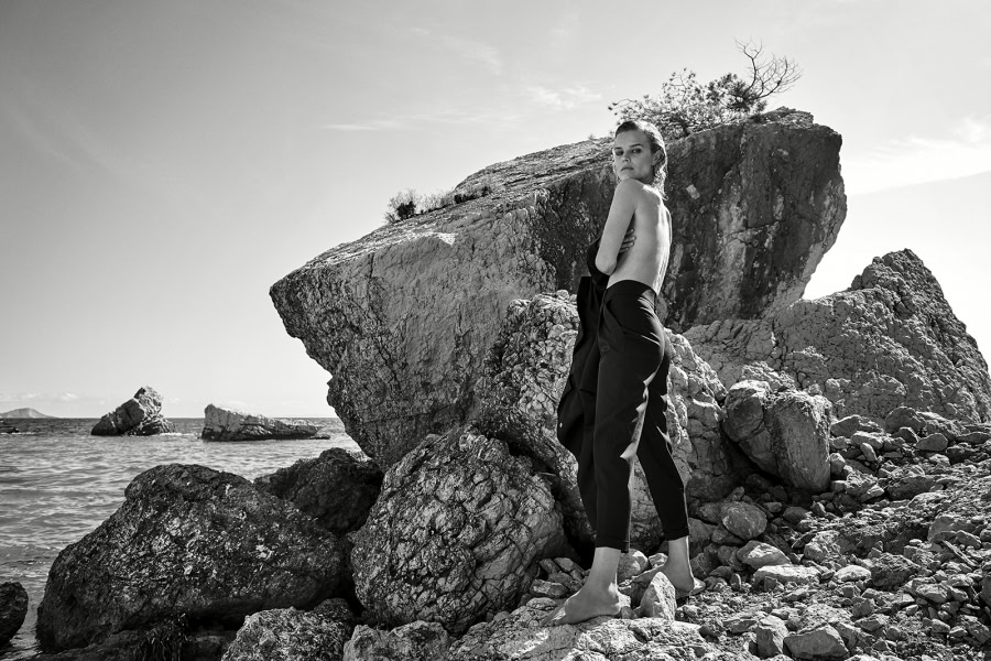 Rugged seaside rock formation with a confident woman standing barefoot, black and white photography, natural landscape, coastal scene, artistic fashion portrait, outdoor photoshoot, free-spirited style, timeless aesthetic.
