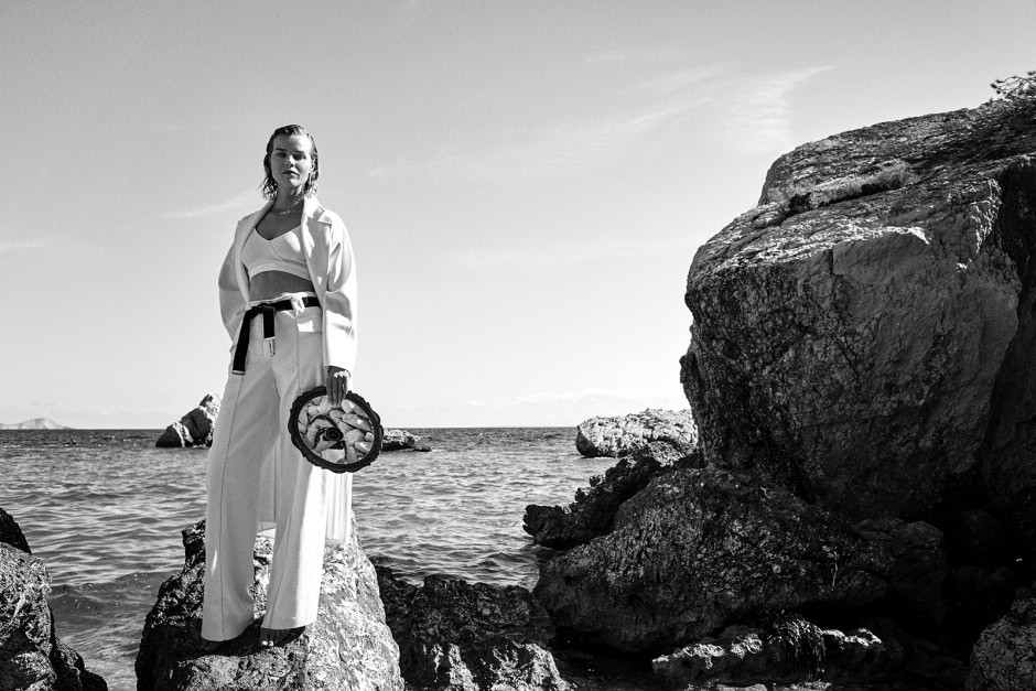 Silhouette of a woman standing on rocks at the beach, holding a round bag, in a black and white artistic fashion photography shot.