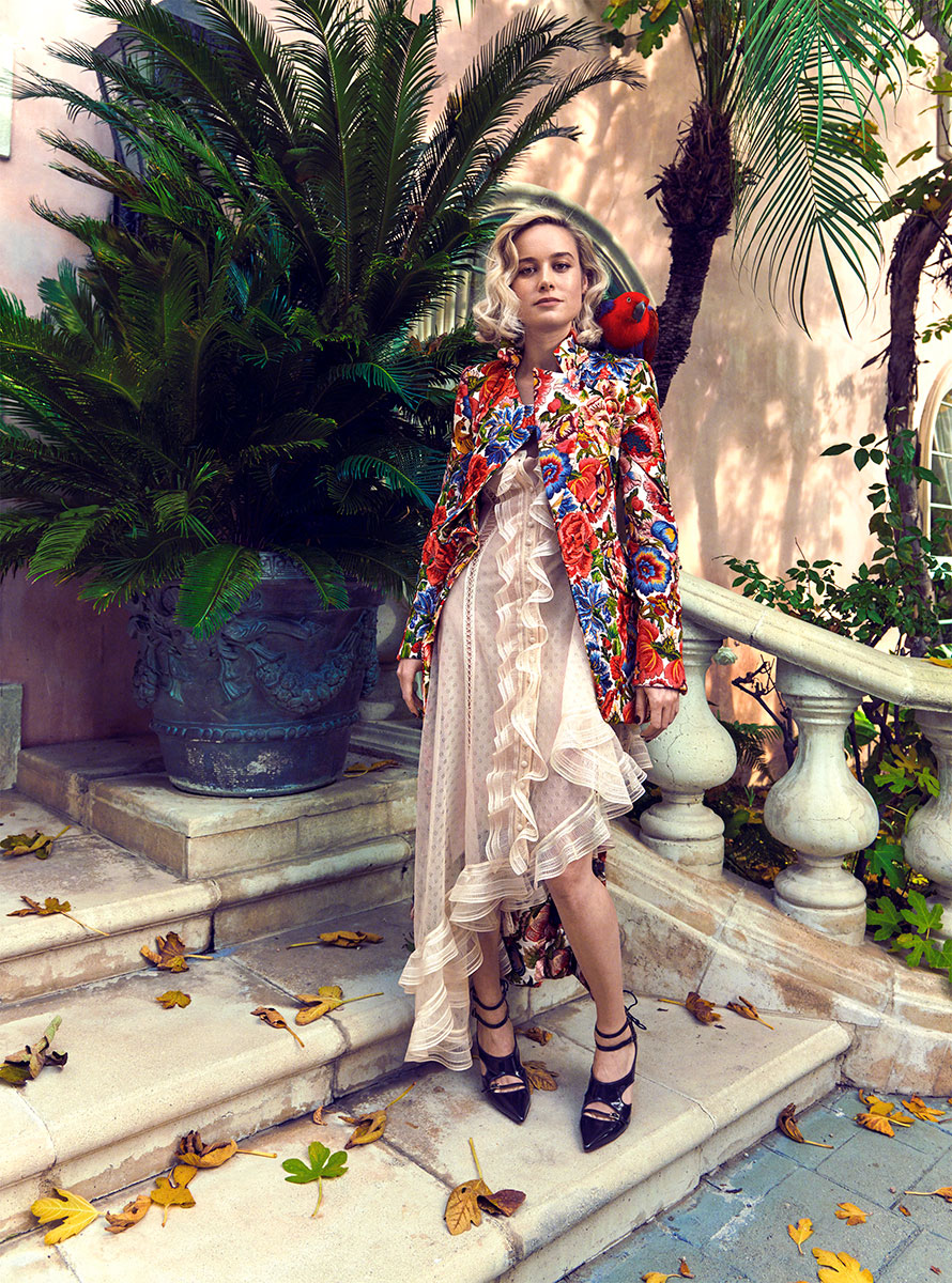 Elegant fashion photography of a woman in a colorful floral jacket and ruffled dress, standing on marble stairs surrounded by lush tropical plants, showcasing high-end fashion and sophisticated style.