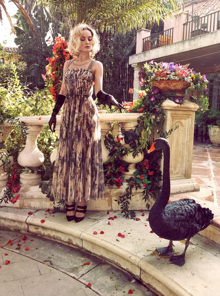 Elegant model in floral dress with black gloves posing outdoors in lush garden setting with swan sculpture and vibrant flowers, showcasing Pamela Hanson Photography.