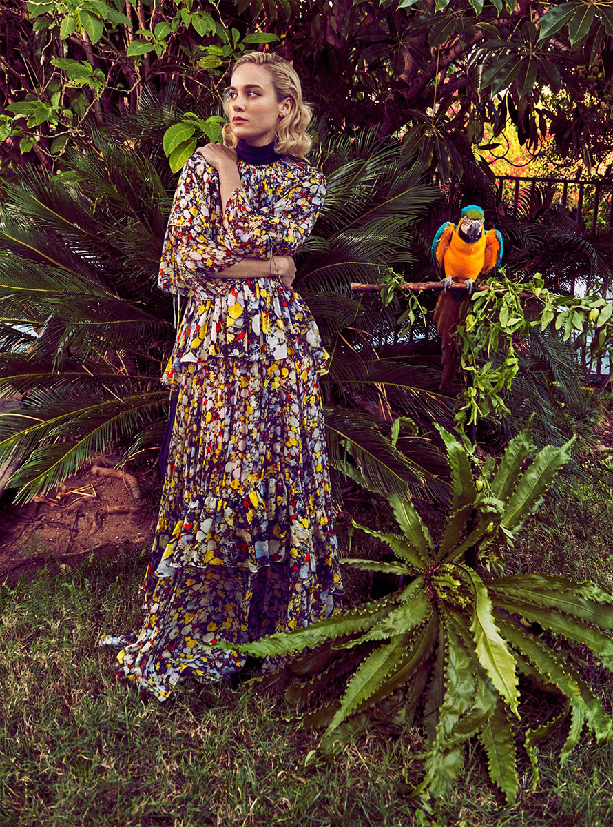 Elegant female model in a floral dress amidst lush tropical foliage with colorful parrot perched nearby.