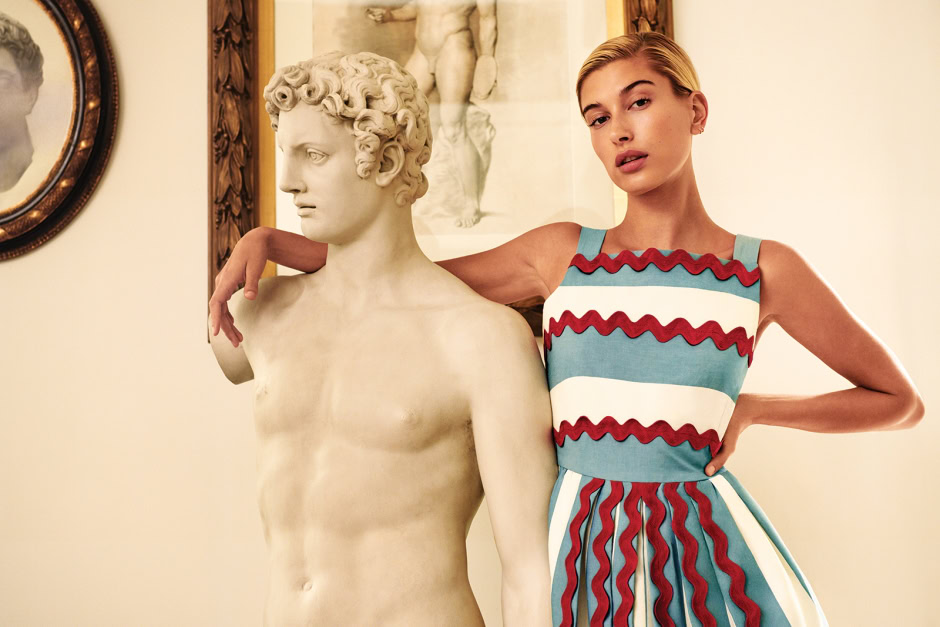 Vivid image of a female model with short blonde hair in a colorful striped dress posing next to a classical marble statue in an elegant gallery setting, highlighting artistic photography and fine art portraiture.