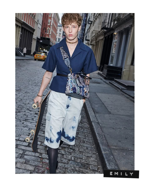 Vibrant street fashion photo featuring a young woman with short hair wearing a blue top, white dyed shorts, black leggings, accessorized with a patterned handbag, skateboard, and city street background, captured by Pamela Hanson Photography.