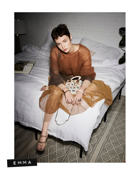 Luxurious fashion portrait of a young woman with short dark hair and bold eyebrows sitting on a white bed, wearing a cozy brown knit sweater, elegant accessories, and a tan layered skirt, in a modern, stylish interior.