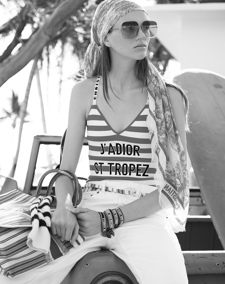 Striped swimsuit with "J'ADIOR ST TROPEZ" text, accessorized with sunglasses, headscarf, and bracelets, exudes a chic, summery fashion vibe at a beach or resort setting.