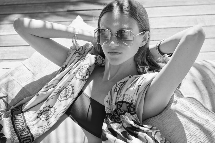 Luxurious black and white portrait of a stylish woman lounging outdoors, wearing oversized sunglasses, a patterned scarf, and a swimsuit, showcasing high-end fashion photography skills by Pamela Hanson.