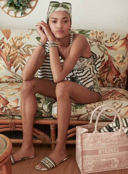 Relaxed woman sitting on floral-patterned sofa in casual summer fashion, featuring striped top, sandals, and accessories, captured by Pamela Hanson Photography.