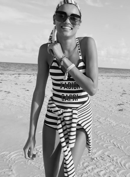 Vibrant beach portrait of smiling woman wearing stylish striped swimsuit, sunglasses, and accessories, enjoying sunny day on sandy shoreline for beach photography, fashion photography, lifestyle photography.